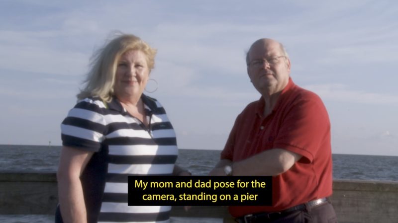 A white woman with shoulder-length blonde hair stands next to a bald man wearing glasses, in front of an ocean and a cloudy blue sky. They both stare at the camera. A caption at the bottom of the image reads: "My mom and dad pose for the camera, standing on a pier".