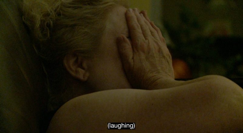 In a dimly lit room, a white woman with blonde hair covers her face with her hands. Her eyes appear to be closed tightly; it's unclear if she's laughing or crying. At the bottom of the screen, an open caption reads: "laughing."