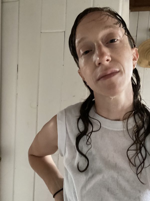  A white nonbinary person with long wet brown hair, wearing a sleeveless white t-shirt, in a sunlit interior 