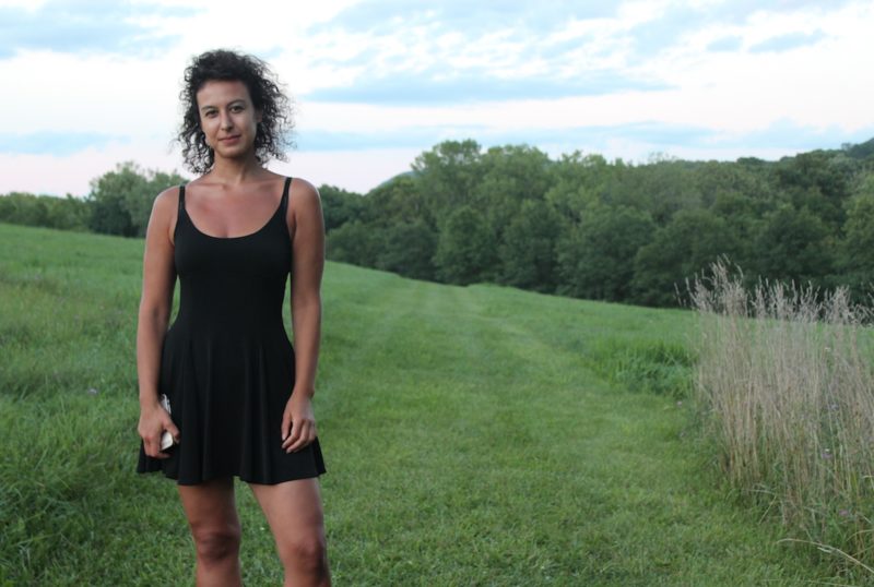 A brown-skinned woman stands toward the left of the frame wearing a fitted black sleeveless dress that cuts mid-way across her thighs. She has black curly hair and looks at the camera with a slight smirk. Behind her is a landscape with a sloping hill covered in lush green grass and a bright blue sky dotted with fluffy white clouds. 