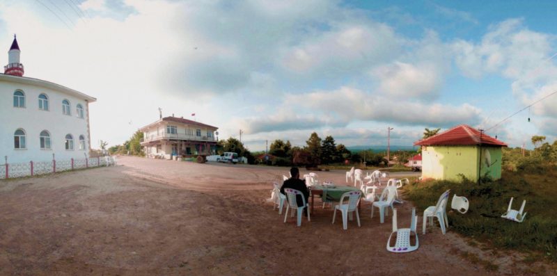 A wide image taken outdoors on a partially sunny day. A dirt road, plastic chairs, and some houses are visible. Someone is sitting on a chair, their back turned against us. On the left, a minaret is visible.