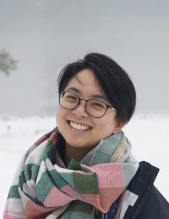 A close up of Elenie Chung, a young woman of East Asian descent wearing glasses and with short black hair. She is wearing a green and pink checkered wool scarf.