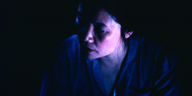 A still from Trinh T. Minh-ha's Surname Viet Given Name Nam. A mostly dark image, with a barely lit woman with black hair looking to the left of the image.