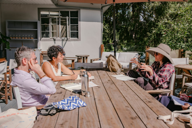 Jason Livingston, Phoebe Cohen, and Caroline Doherty sitting outside on a wooden table at a retreat hosted by the Simons Foundation in September 2023. It is a sunny day; Caroline Doherty is speaking.