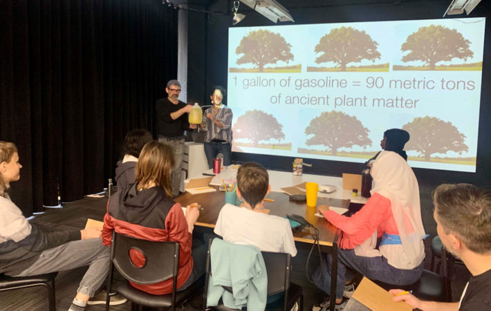 Jason Livingston, Phoebe Cohen are standing, holding a gallon bottle of gasoline. Several students are gathered around a large table with notebooks. Behind Livingston and Cohen is a projected slide with trees and the words “1 gallon of gasoline = 90 metric tons of ancient plant matter”
