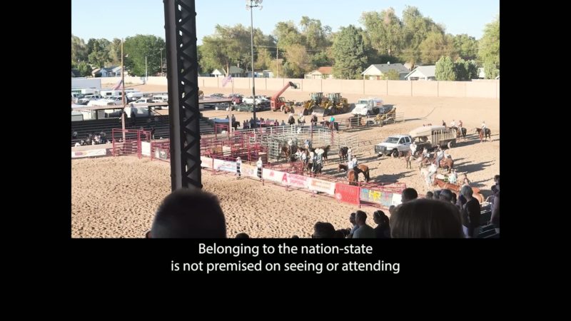 A still from the film An All-Around Feel Good. An image of the Colorado State Fair, taken from the bleachers on a bright sunny day. The camera is facing a holding pen, where there are horses and riders, numerous parked cars and a tractor, and several U.S. flags, and audience members both near the camera and across the arena. The image is surrounded by a black border on both sideas and at the bottom. In the blank space at the bottom is a caption that reads: “Belonging to the nation-state is not premised on seeing or attending.”