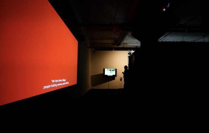 A view of the installation of Jordan Lord’s exhibition “The Voice of Democracy” in Squeaky Wheel’s gallery space. On the left is I Didn’t Set Out to Make a Film About Religion, a red projected image on a wall, with a red color field and the captions “‘We Heart New College”, people holding cameras, police officers”. On the far end of the room is a person looking at a dimly lit television screen wearing headphones, featuring An All-Around Feel Good. On the television are a tree and captions underneath.