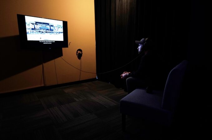 A person wearing a hat and sitting on a couch, watching a television screen that is showing An All-Around Feel Good. He is wearing headphones. An arena is on the screen, along with the caption “This barrier quite literally marks national borders”.