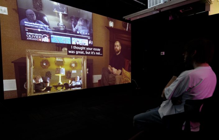 A seated person viewing How Is It That You Frame Old Glory In Your Mouth?, a video projected on a screen. On the screen is Jordan seated with their arms and legs crossed, and one eyebrow slightly raised. They are looking towards the camera. Next to them is a mirror showing Lord’s parents, Deborah and Albert, who are speaking to Jordan. Above the mirror is a television screen with video of Jordan’s parents as it's been paused. There are captions in the middle of the screen: “I thought your essay was great, but it’s not…”