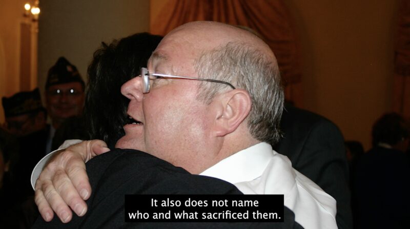 A still from How Is It That You Frame Old Glory In Your Mouth?. Jordan and their father Albert are hugging in a photograph from the mid-2000s, taken by their mother Deborah. Albert’s face mostly fills the frame, as they hug in profile to the camera. He looks ahead with his mouth open, as he tells Jordan something. It might be a smile or he might be tearing up. It's hard to tell because of his glasses. A caption on the bottom of the still says "It also does not name who and what sacrificed them."