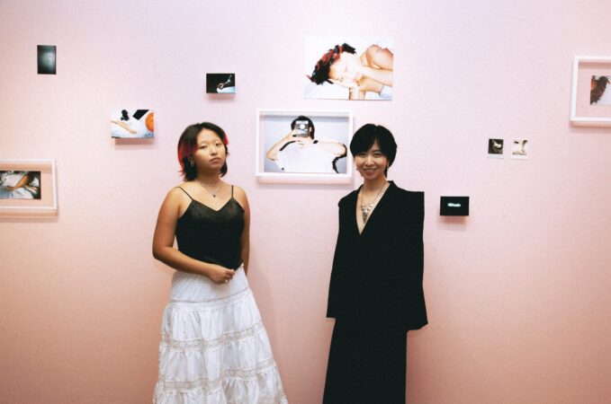 huiyin zhou in black top and white skirt on the left and Laura Dudu with black long dress on the right, both standing in front of a pink wall of pictures and frames in various sizes. 
