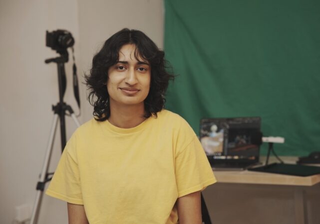 Ajunie Virk sitting in front of camera equipment, a laptop, and a green screen.
