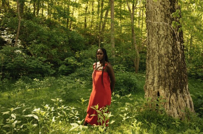 Arielle Knight in a red dress in a green sun-spackled field with trees and bushes.