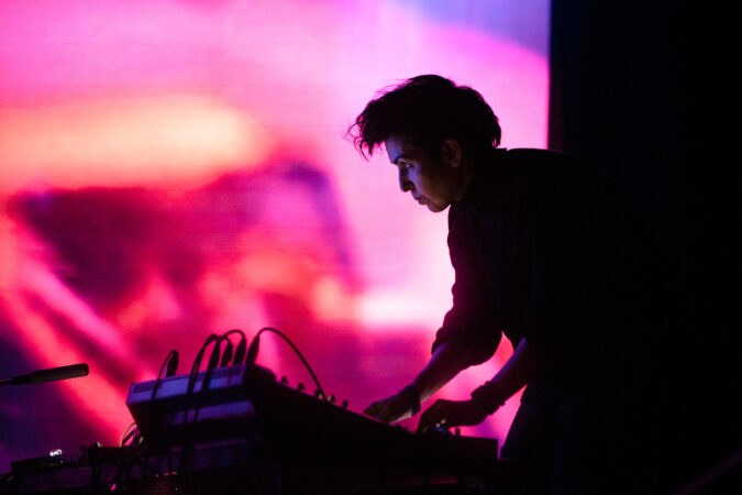 A sound artist performing on stage in silhouette, leaning over a mixer and electronics, backlit by a vivid pink and orange projected backdrop.