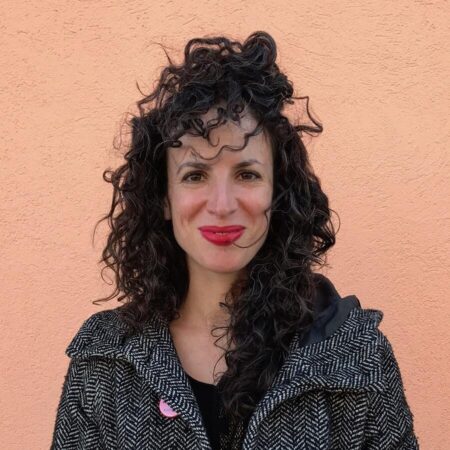 A curly haired woman with red lipstick and a peacoat, who appears to be in her early 30s but is now a decade older. 