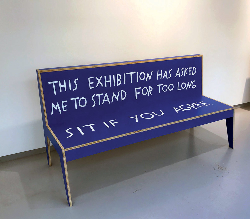 A blue bench with hand-painted white text reads: This exhibition has asked me to stand for too long. Sit if you agree.