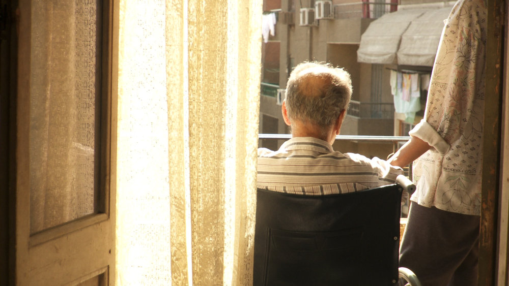 A seated man on a balcony with balding hair with his back turned against us, with a woman's hand on his shoulder. Bright sunlight is reflected off a curtain.