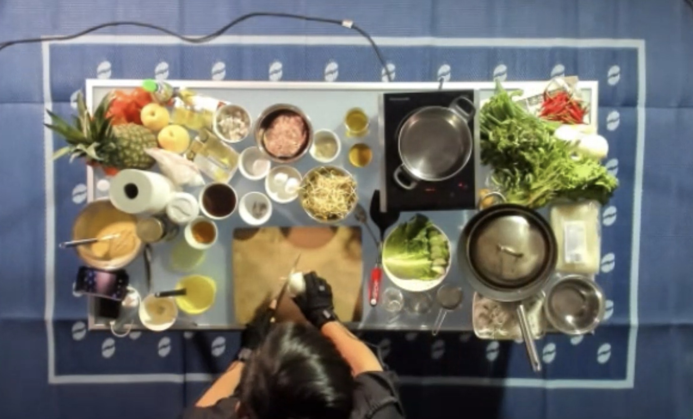 An overhead photograph of a table with several dishes of ingredients and plates. A man on the bottom half of the image is cutting an onion while wearing motion capture gloves.