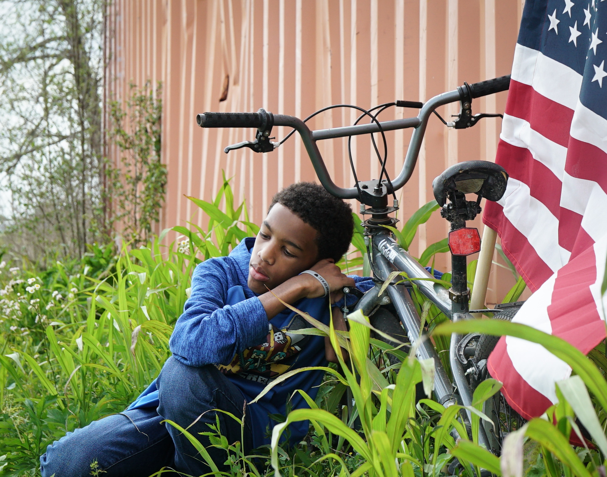 Muse Dodd, Kendi (2018). A young Black child rests their head, eyes closed on their bike in a field of overgrown grass. The American flag is propped against a rust colored storage unit and blows gently in the wind.