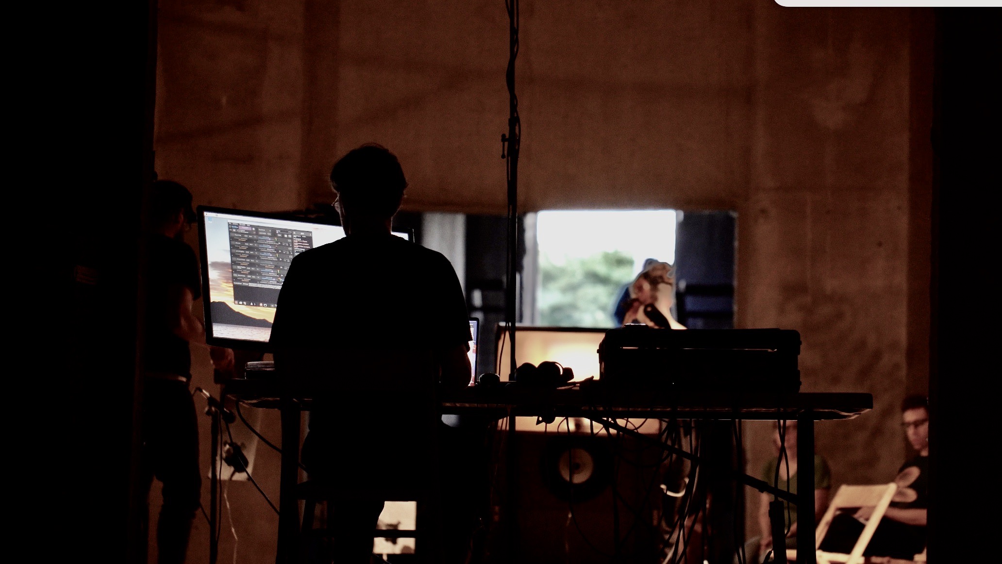 A photograph of Rob Cosgrove as he performs his sound piece, Floaters inside Silo City in September 2022. Rob is seen from behind inside a large concrete structure. He is facing a computer. In the background, you can see another musician performing as part of the work.