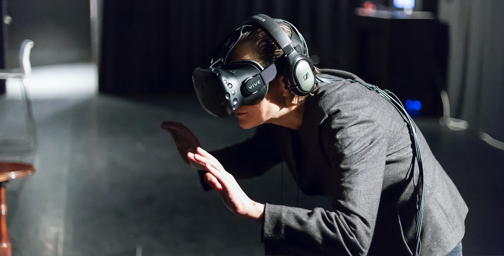 Documentation of an installation of Deniz Tortum, September 1955 (2016). A person wearing virtual reality goggles in a sunny room. They are crouched and their hands are facing out as if they are gently pushing something.