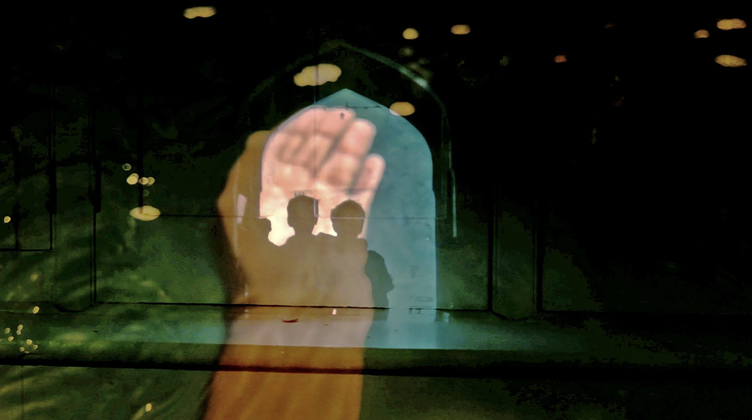 Kalpana Subramanian, Incantation (2021). A photograph depicting multiple images projected on a surface. The shadows of what look like two young people, arms on each other's shoulders, standing under an arched gateway to a historic building in Delhi, India. Upon this image is an extended hand, palm open. It looks like the young people are in the palm.
