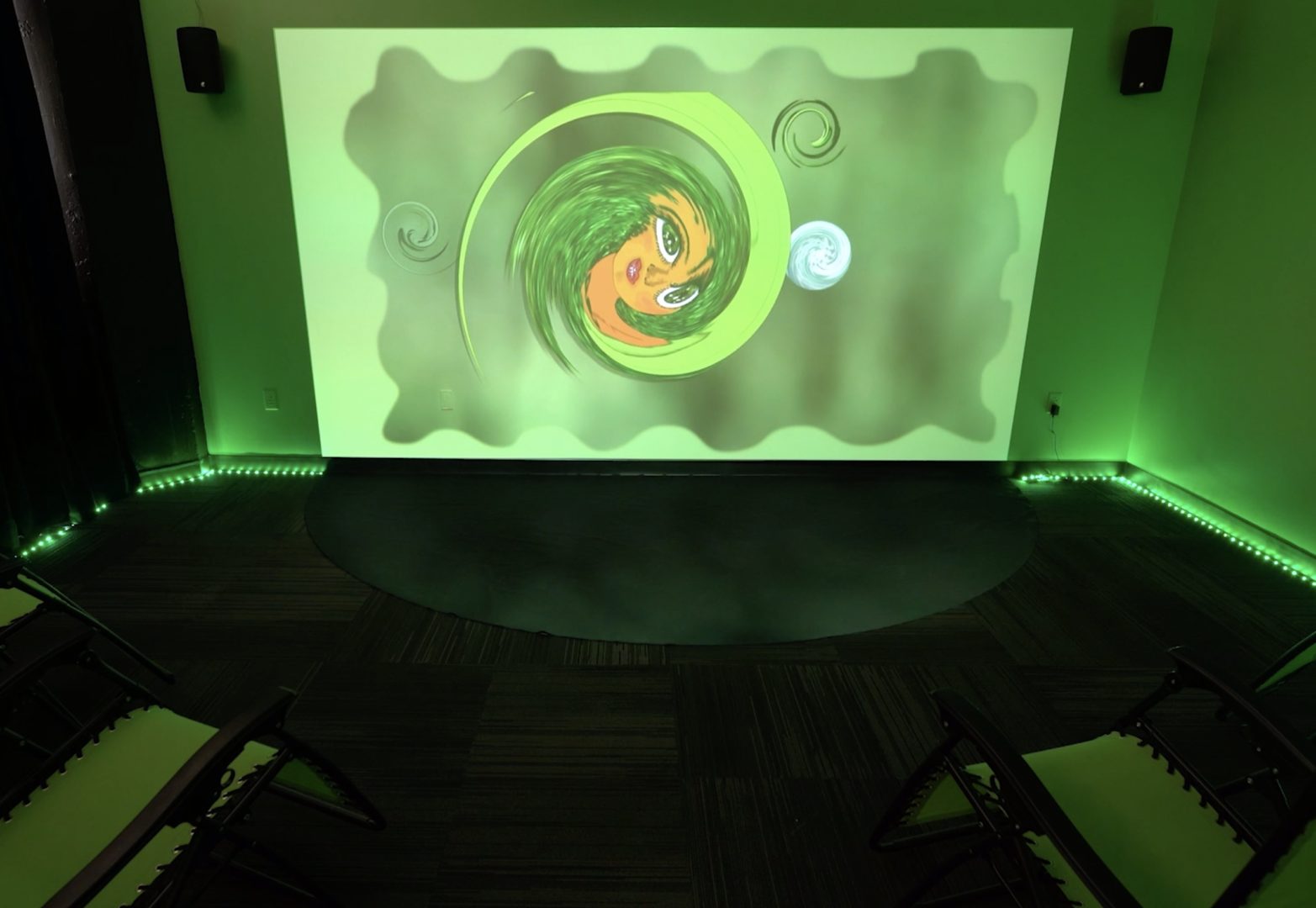 Documentation of hiba ali, oceans we carry: rough as silk. The video in the cyclone (rough as silk) is projected on a wall; a green wavelike video is projected on a canvas on the floor. There are two green chairs facing the wall.