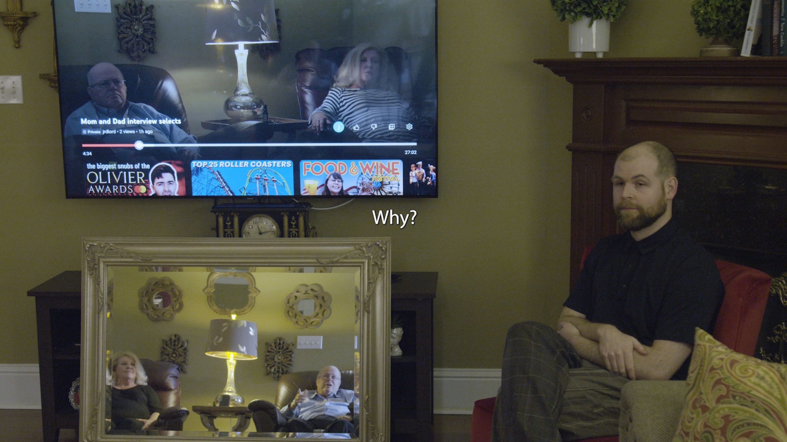 A still from the film Voice of Democracy shows a dim living room, in which a tv hangs on the wall. The tv displays two older white people, the filmmaker's parents, sitting with a gold lamp in between them. The filmmaker's father looks angry and their mother looks concerned. The tv image is paused on a Youtube window, which reads: "Mom and Dad interview selects," below which a series of other videos are recommended including "The Biggest Snubs of the Olivier Awards," "Top 25 Roller Coasters," and "Food & Wine Festival." Reflected in a mirror below the television are the filmmaker's parents sitting in the same set-up as their image on the television, with the same gold lamp between them. They are looking across the room at the filmmaker, a 30-something white person with a buzzcut and a beard, who looks back at them with their arms folded and legs crossed, with a serious and sympathetic look. Their father is pointing at the television, saying something. In the center of the image, between the mirror and the television, is a caption that reads, "Why?"