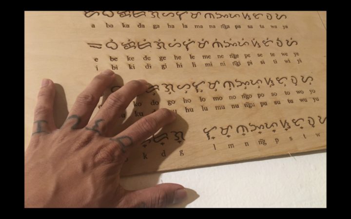 A still from english is yr mother tongue by champoy. A hand with tattoos is tracing over a paper with translations between languages.