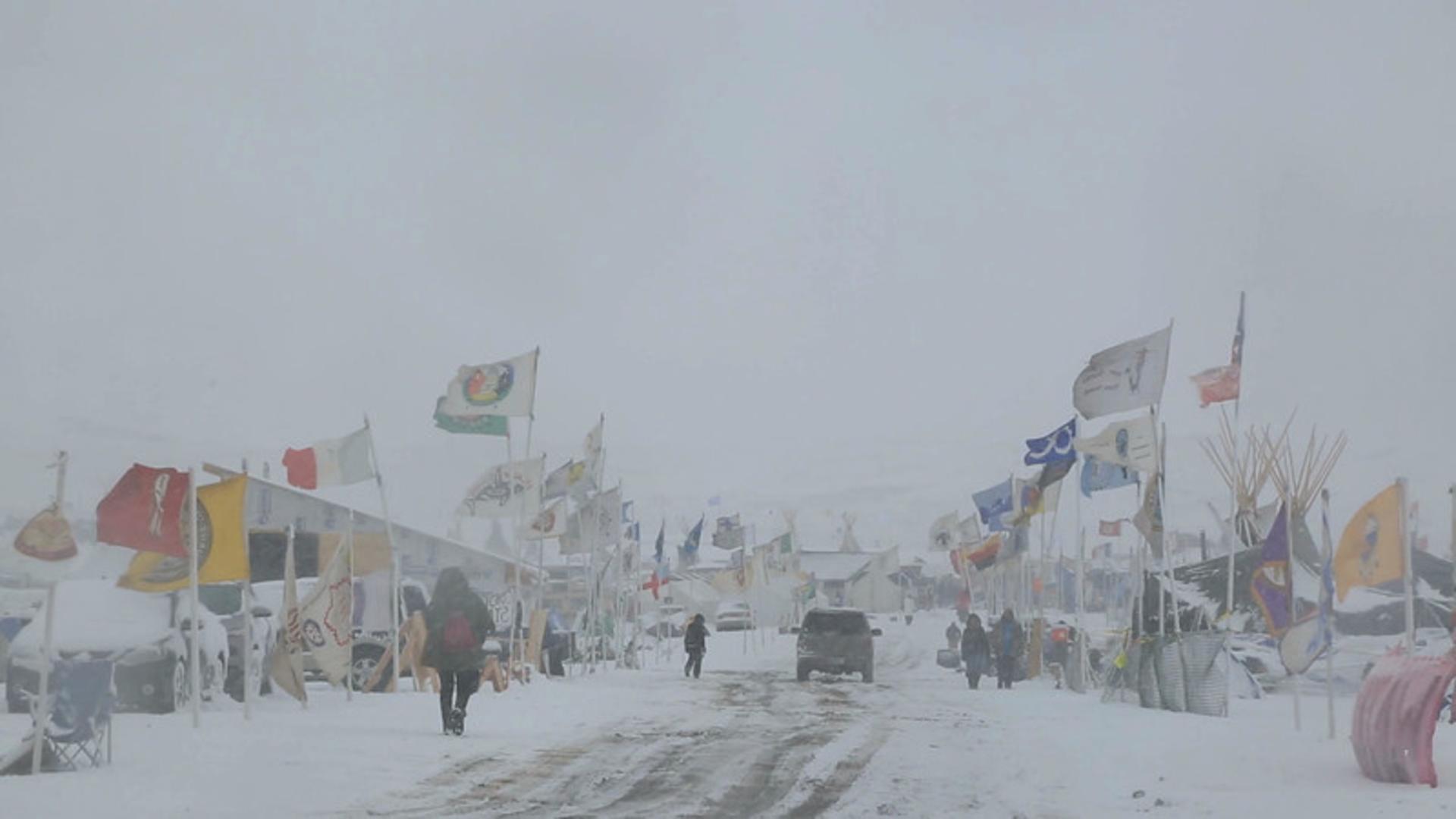 A still from the film Spaces of Exception, showing Standing Rock covered in snow under a gray sky. There is a road with a car in the middle of the frame, and a few people in big coats. Over 50 flags of Indigenous nations and other countries are on either side of the road.