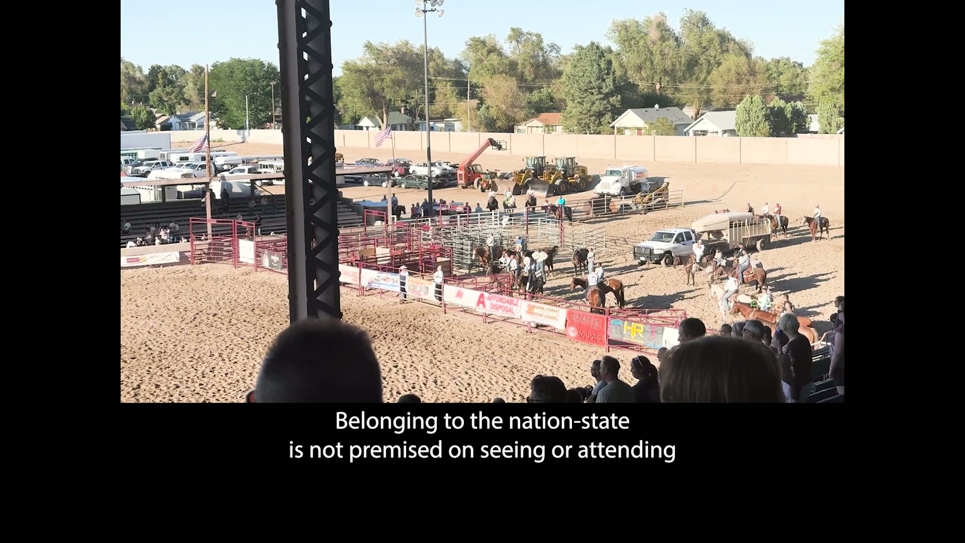 A still from the film An All-Around Feel Good by Jordan Lord. Prominent black borders are on the left, right and bottom edges of the image.  Framed by the black borders is an image of the Colorado State Fair, taken from the bleachers on a bright sunny day. The camera is facing outside the main stage, where there are horses and riders in a pen, numerous parked cars and a tractor, and several U.S. flags, and audience members both near the camera and across the stage. On the bottom is the caption "Belonging to the nation-state is not premised on seeing or attending".