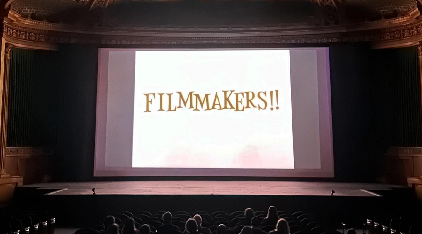 Documentation from the 20th Animation Fest retrospective at North Park Theater. A projection in a darkened movie theater. On the screen is the word "Filmmakers!", which is from Helen Hill's 2004 short film "Madame Winger Makes a Film: A Survival Guide for the 21st Century"