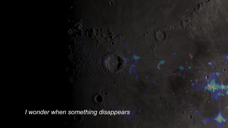 A still from Noor Abuarafeh's The Moon is a Sun Returning as a Ghost. An image of the moon in grey, black, and patches of purple blue, with the words "I wonder when something disappears" on the bottom left.