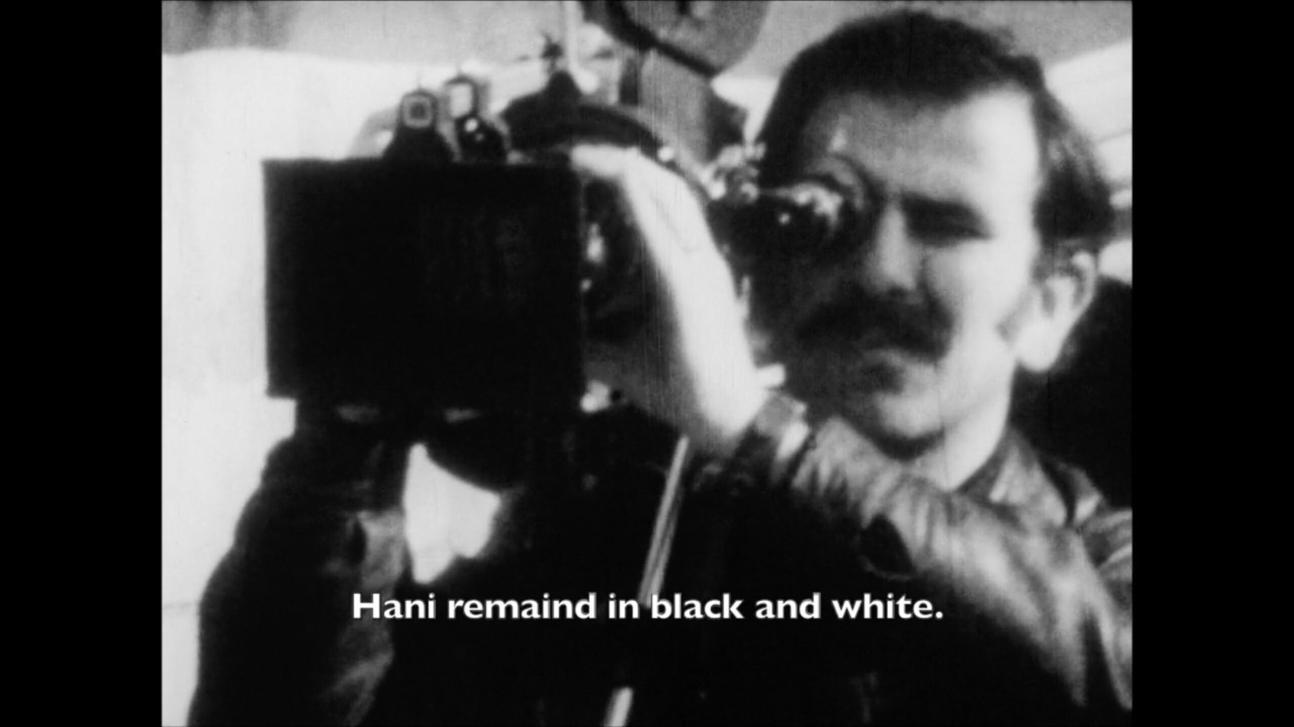 A still from Azza El-Hassan's A Remake of a Revolutionary Film. A photograph of the Hani Jawharieh holding a 16mm camera in black and white. On the image is the subtitle, "Hani remained in black and white."