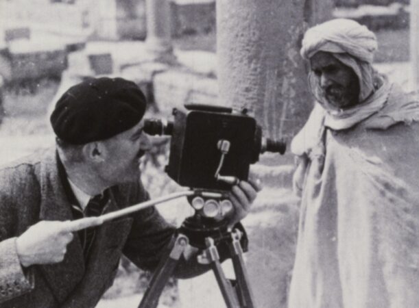 A still from Assia Djebar's The Zerda and the Songs of Forgetting. A French person shooting with a 16mm camera. An Algerian man wearing a burnous is to his right, looking at him. The French mans gaze and lens is past the Algerian.