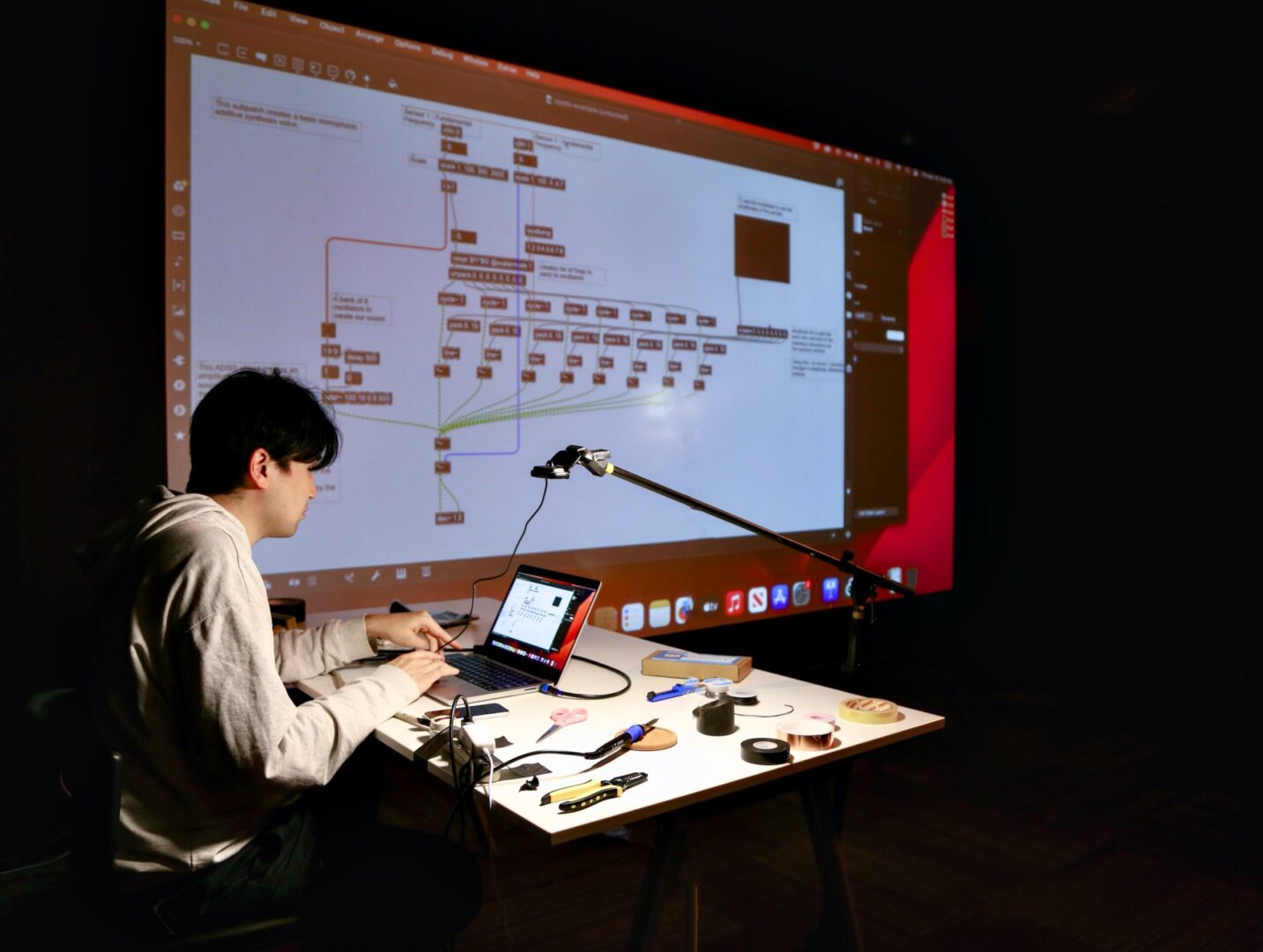 The artist Jaehoon Choi, surrounded by various electronic equipment and soldering iron. Behind him is a projector showing the connection of various signals.