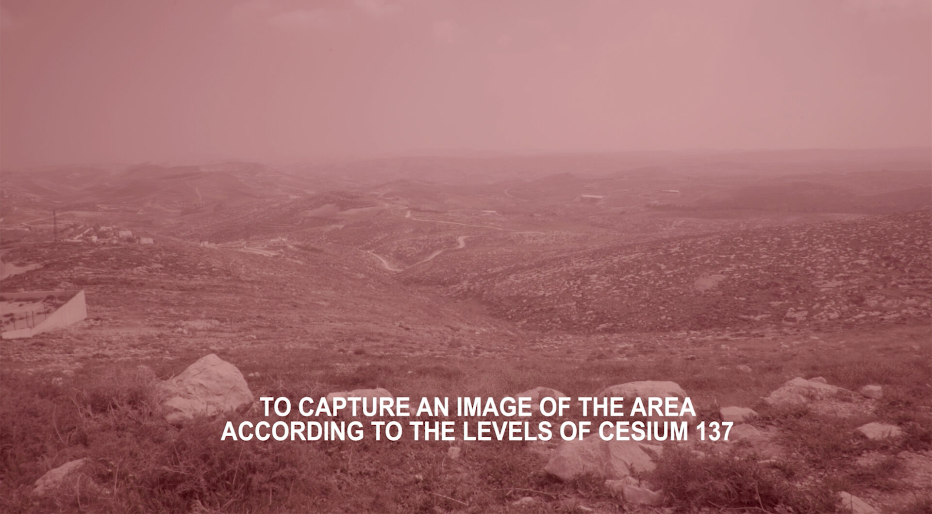 Still of Inas Halabi, We Have Always Known the Wind’s Direction (2019). A red and pink tinted filtered image of a Palestinian landscape with white subtitles. The subtitles state "TO CAPTURE AN IMAGE OF THE AREA ACCORDING TO THE LEVELS OF CESIUM 137".