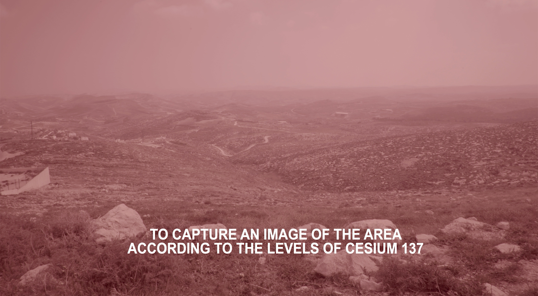Still of Inas Halabi, We Have Always Known the Wind’s Direction (2019). A red and pink tinted filtered image of a Palestinian landscape with white subtitles. The subtitles state "TO CAPTURE AN IMAGE OF THE AREA ACCORDING TO THE LEVELS OF CESIUM 137".