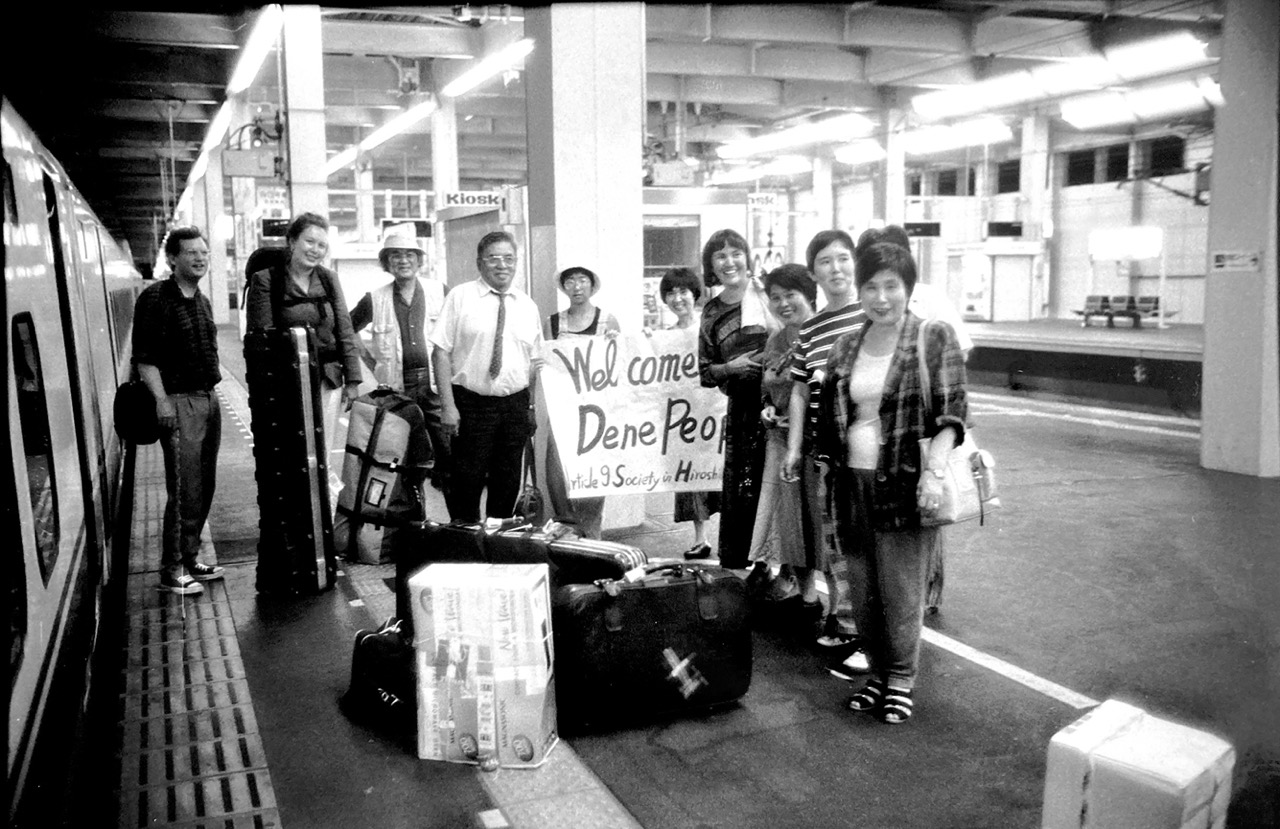 A photograph from the production of A Village of Widows. A number of people are standing around a train station in Japan with luggage around them. Two of them are holding a sign "Welcome Dene People" and underneath "Article 9 Society in Hiroshima". Photograph courtesy of Peter Blow.