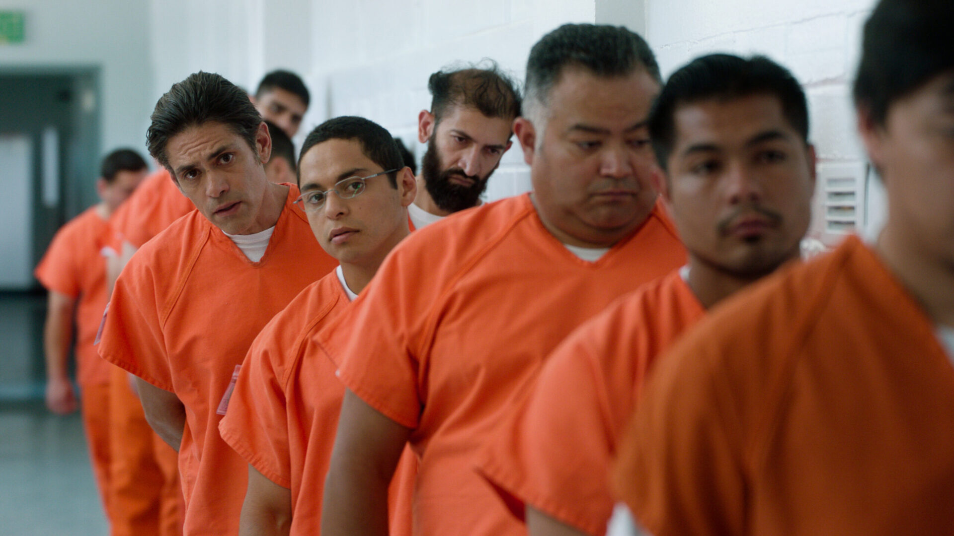 A still from the 2019 film The Infiltrators. A row of detainees in orange jumpsuits. Two people are clearly in focus.