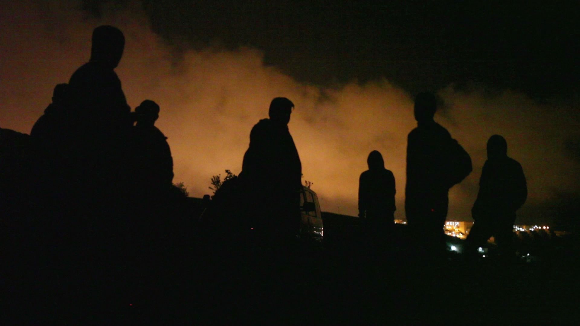 A still from Khaled Jarrar's film, Infiltrators. The frames of several people can be discerned against a city backdrop at night time. Clouds are lit in orange from the city lights.