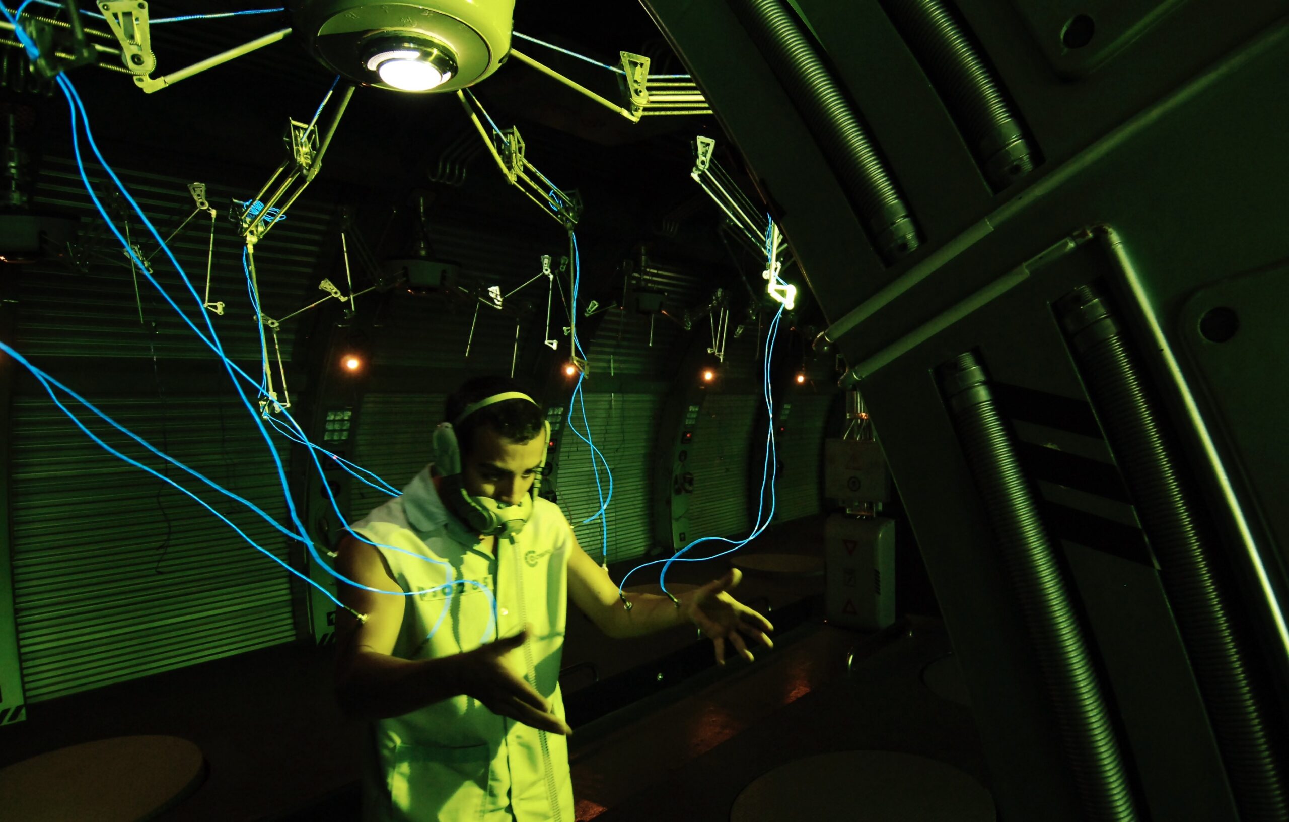 A still from Alex Rivera's film Sleep Dealer. A man with his mouth obstructed by some strange technology is connected to a larger machine by a mess of blue wires. He is looking intently ahead as if looking somewhere else.