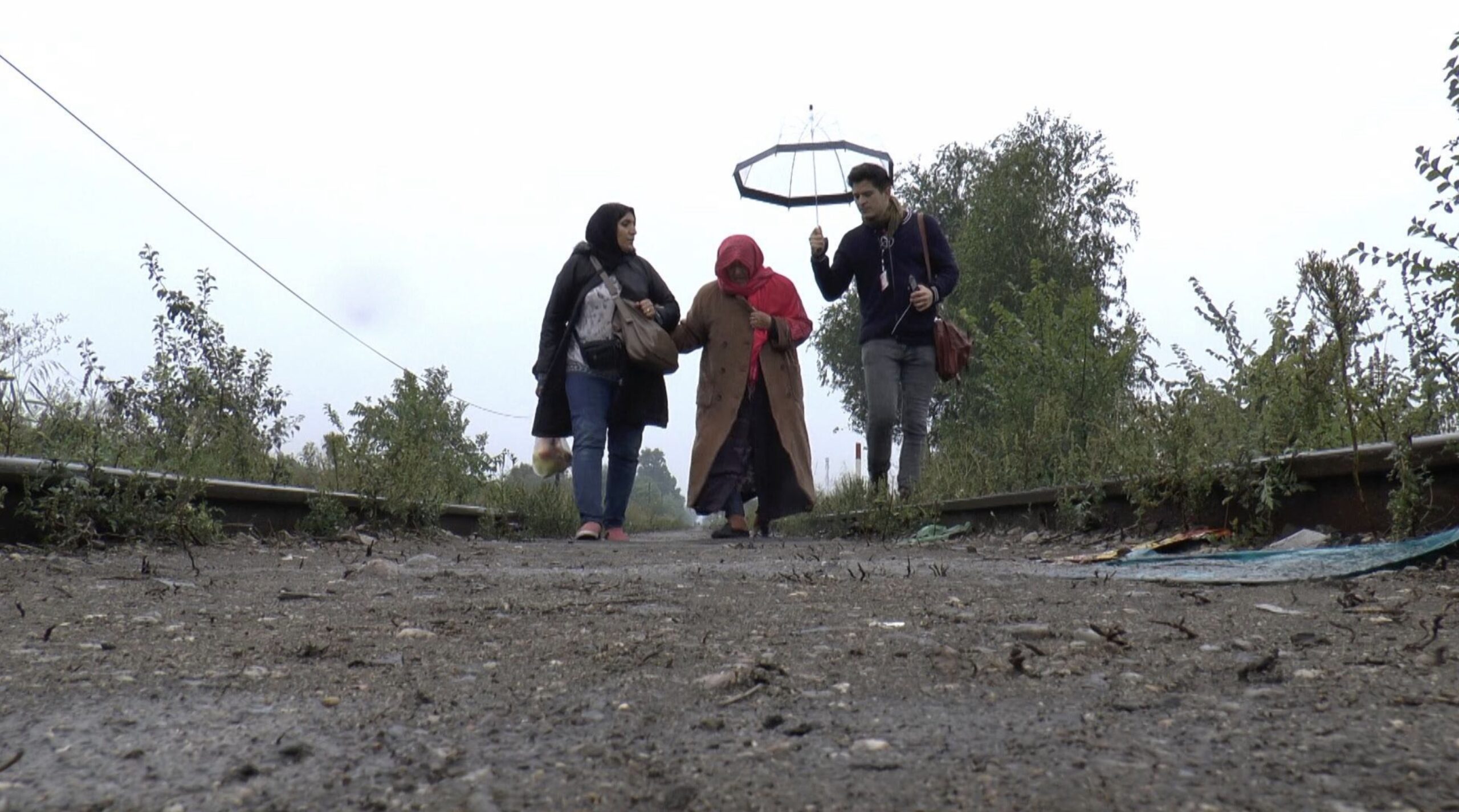 A still from Khaled Jarrar's film, Notes on Displacement. A man and two women are walking along train tracks on a cloudy day. The man is carrying an umbrella. One of the women has multiple bags hanging from her shoulder.