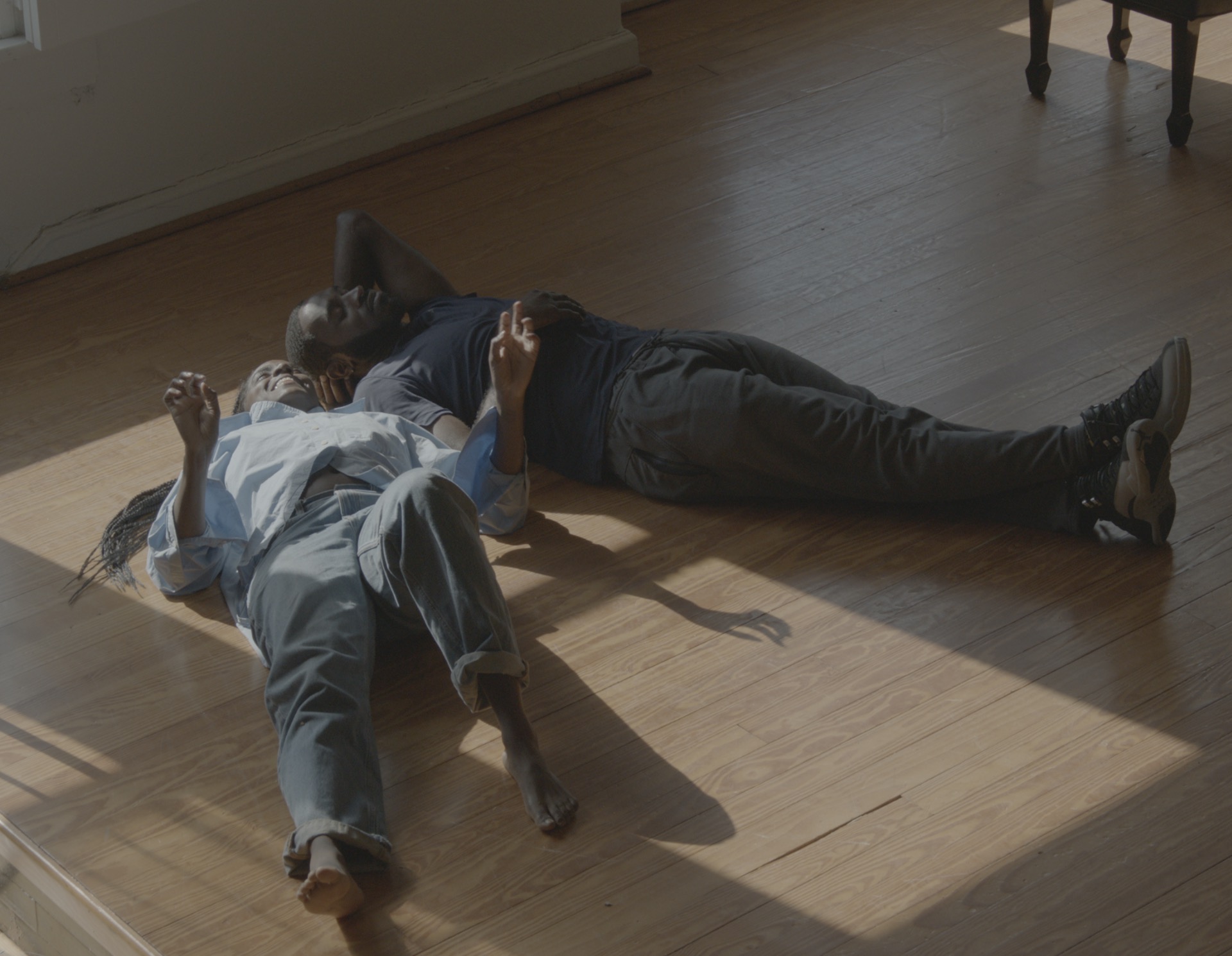 A still from And Counting a film by Arielle Knight. Two Black people laying on a wooden floor, their heads next to each other. One of them is smiling while gesturing and talking, the other has their eyes closed, their head resting on their hands. The sun beams on them from a window.