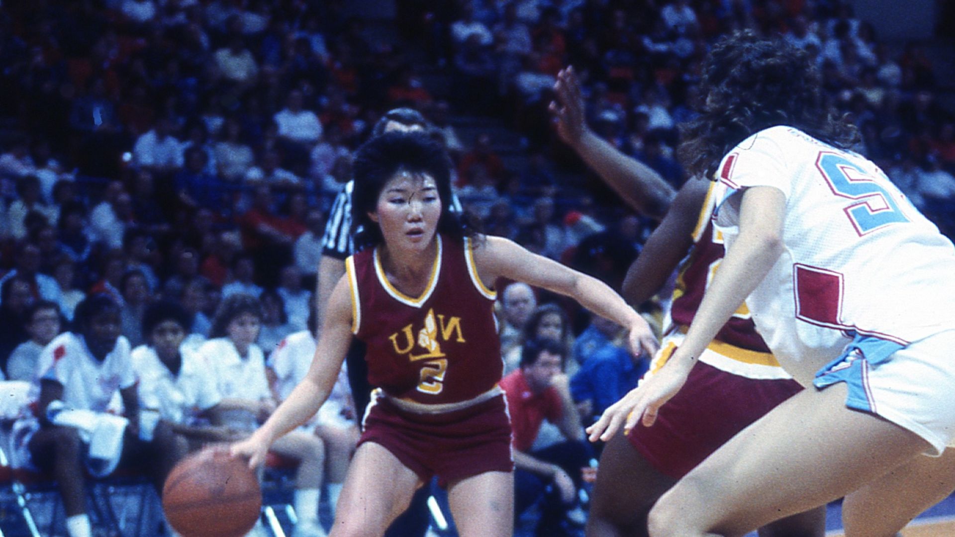 A petit young Asian woman wearing a burgundy red jersey and dribbling a basketball in front of an opposing tall player in a white jersey, all within the backdrop of a crowded arena.