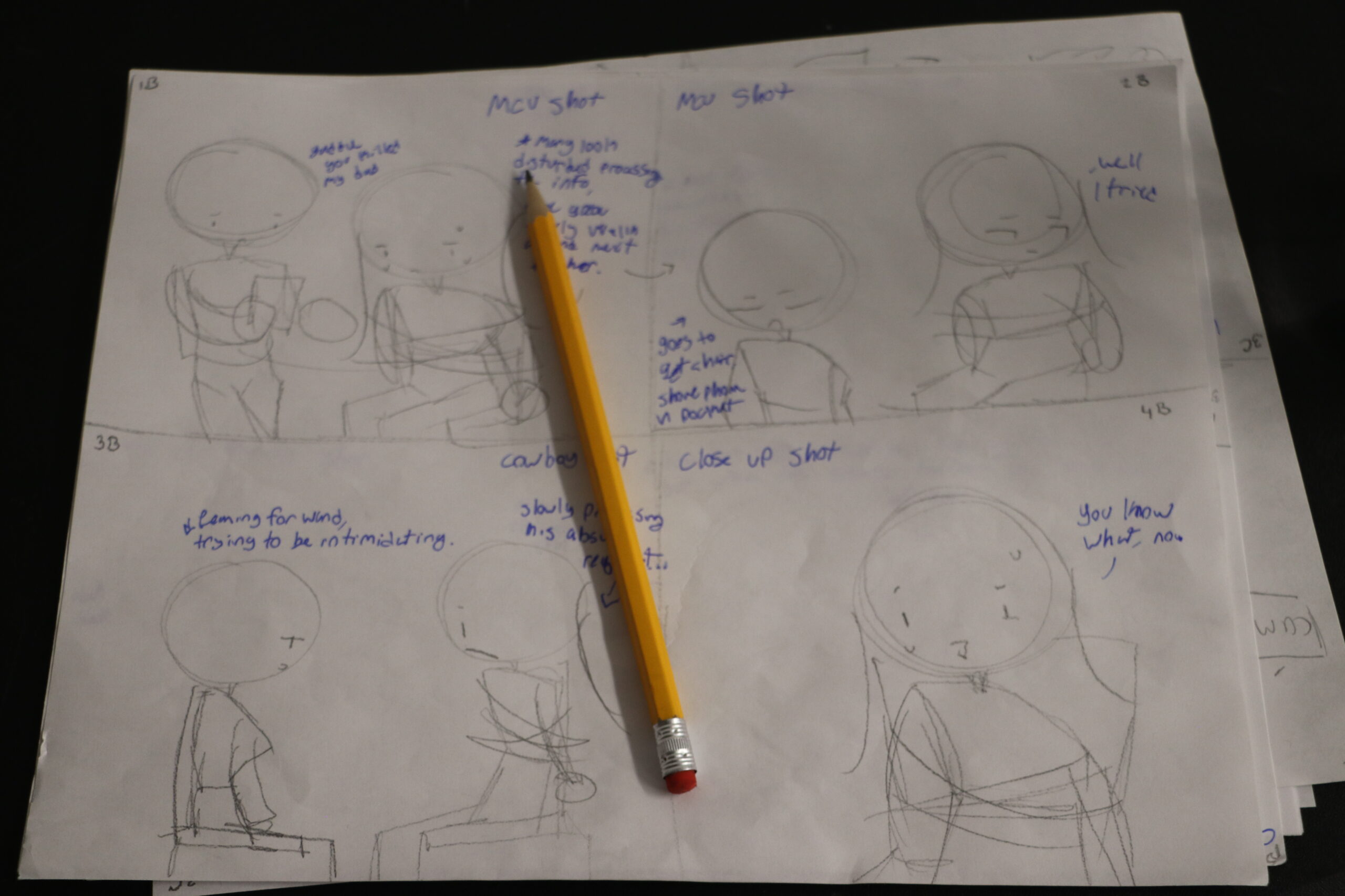 A 4-panel storyboard showing simply drawn figures and dialogue. A wooden pencil lies on top of the paper.